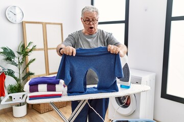 Senior caucasian man ironing holding burned iron shirt at laundry room in shock face, looking skeptical and sarcastic, surprised with open mouth