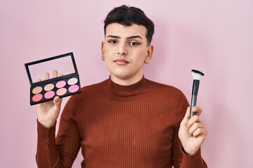 Non binary person holding makeup brush and blush relaxed with serious expression on face. simple and natural looking at the camera.