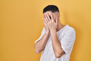 Young handsome man wearing casual t shirt over yellow background with sad expression covering face...