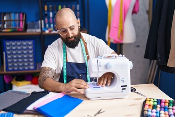 Young bald man tailor smiling confident using sewing machine at clothing factory