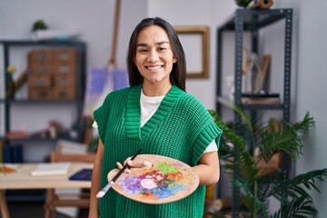 Young south asian woman holding painter palette looking positive and happy standing and smiling with a confident smile showing teeth