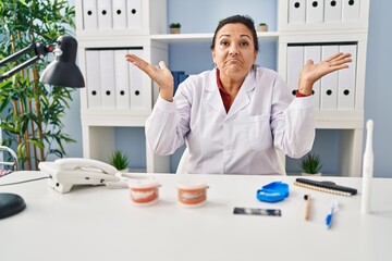 Hispanic mature dentist woman working at the clinic clueless and confused expression with arms and...