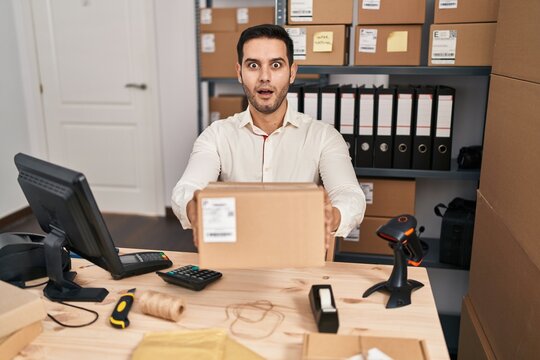 Young hispanic man with beard working at small business ecommerce holding box afraid and shocked with surprise and amazed expression, fear and excited face.