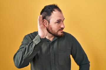 Fototapeta premium Plus size hispanic man with beard standing over yellow background smiling with hand over ear listening an hearing to rumor or gossip. deafness concept.