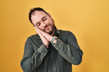 Plus size hispanic man with beard standing over yellow background sleeping tired dreaming and posing with hands together while smiling with closed eyes.