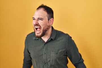 Plus size hispanic man with beard standing over yellow background angry and mad screaming frustrated and furious, shouting with anger. rage and aggressive concept.