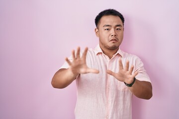 Chinese young man standing over pink background afraid and terrified with fear expression stop gesture with hands, shouting in shock. panic concept.