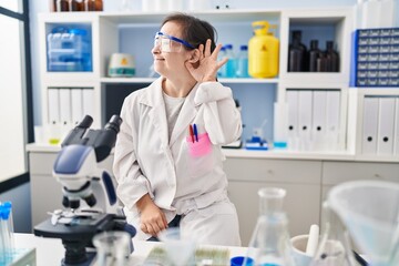 Hispanic girl with down syndrome working at scientist laboratory smiling with hand over ear listening an hearing to rumor or gossip. deafness concept.