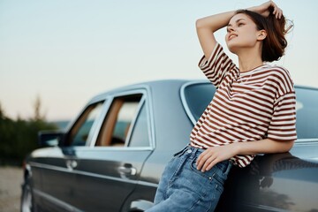 A young woman in a striped T-shirt and jeans stands by her car relaxing from driving, leaning on her car beside the road and smiling. travel as a lifestyle and freedom of movement 