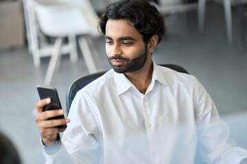 Obraz na płótnie Canvas Indian young business man company worker, professional employee, stock market trader holding smartphone using cell phone mobile apps checking financial market data in application at work in office.