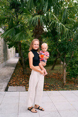 Smiling mother with a little boy in her arms stands under a palm tree in the garden. High quality photo