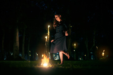 Portrait of beautiful witch in black dress with magic fire ball in forest at dark night