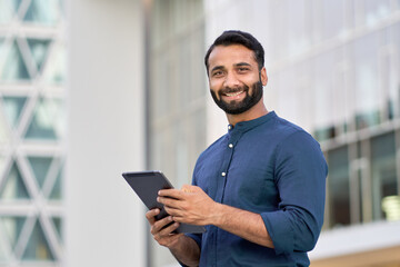 Smiling young adult indian business man professional executive holding digital tablet online fintech outside, happy eastern businessman ceo using pad standing outdoors in big urban city street.
