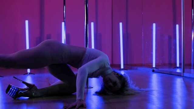 Young woman pole dancer performs acrobatic tricks on pylon indoors. Blonde slender woman dancing near pole with pink and blue light at background. Poledance practice in 4K, UHD