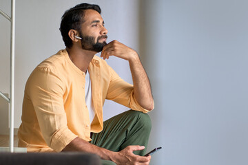 Thoughtful bearded indian man user sitting at home on stairs relaxing wearing earbuds using smartphone thinking while listening to music podcast or audio book in mobile application on cell phone.