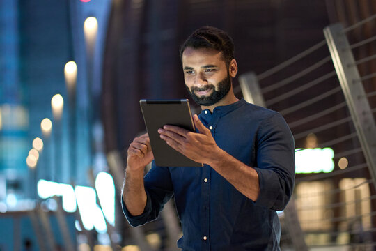 Smiling young indian business man professional, eastern businessman executive standing outdoors on street reading ebook holding using digital tablet online technology in night city with urban lights.