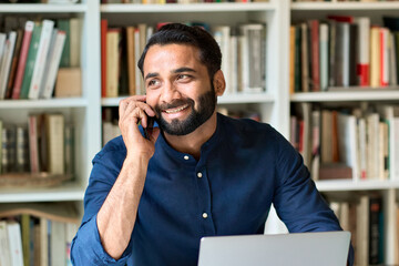 Happy smiling eastern indian professional business man sitting at work desk talking on cell phone,...