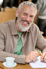 Happy older adult business man professor or student writing in notebook, smiling middle aged gray-haired bearded author or writer taking notes looking at camera sitting at table. Vertical portrait. - obrazy, fototapety, plakaty