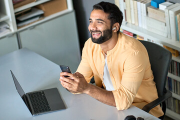 Happy smiling ethnic indian professional business man manager sitting at work desk wearing earbud using cell phone, laughing eastern businessman having virtual mobile chat video call on smartphone.