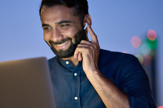 Smiling Indian Business Man Wearing Earbud Having Virtual Meeting Online Remote Conference Video Call Looking At Laptop Computer Remote Working, Watching Webinars Sitting At Night In Office.