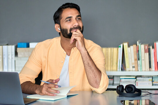 Thoughtful dreamy indian business man writer or student working or learning online, listening podcast looking away thinking of new ideas sitting in office at home using computer, planning project.