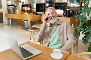 Happy smiling middle aged professional business man, older mature entrepreneur sitting at cafe table talking on cell phone making mobile call remote working online on laptop and having morning coffee.