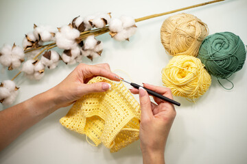Woman's hands are knitting baby clothes from yellow yarn, on a white table, top view.