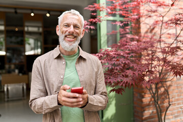 Smiling gray-haired older middle aged bearded man using mobile phone outdoors. Happy old senior...