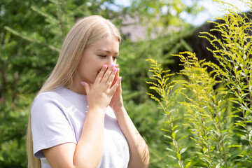 A young blonde girl stands near an ambrosia plant, covers her face with her hands and sneezes. Allergy to seasonal bloom