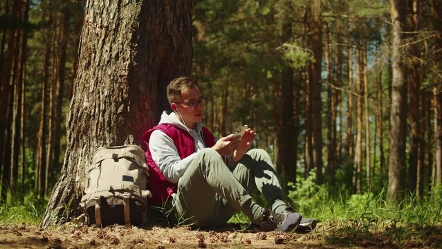 Asian man tourist listening to music, using earphones. Young hiker resting in forest camp, traveling and hiking alone, resting. Summer tourism, wild nature.