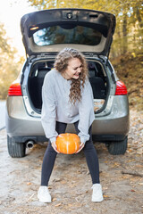 Girl is holding freshly picked organic orange pumpkin near car. Woman is having fun. Autumn season