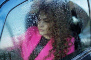 Sad woman looks out foggy car window. Window with rain drops. Curly-haired woman is riding in car