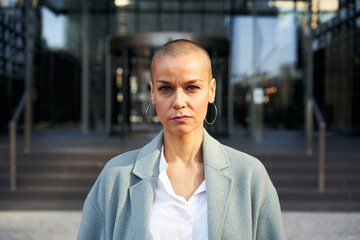 Serious middle-aged empowered woman with shaved hair looking at camera. Business people outdoors on...