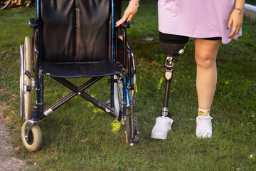 Young woman with amputated leg using prothesis staying in the grass near wheelchair for disabled people 