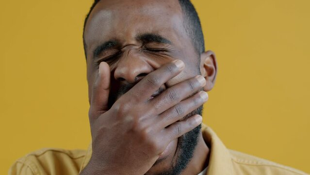 Close-up portrait of tired young man yawning and looking at camera on yellow background. Exhausted people and expressions concept.