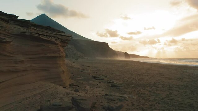 Drone flight over endless sand beach Cofete. Volcanic mountain landscape. Golden sandstones in sunset warm light. Ocean water evaporation. Dust, mist, fog rises above the waves. Fuerteventura Island.