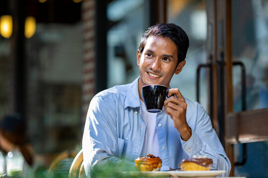 Asian man sipping a hot espresso coffee while sitting outside the european style cafe bistro enjoying slow life with morning vibe at the city square with sweet pastry