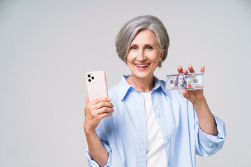 Mature grey haired woman 50s with dollar bill and smartphone in hands showing it on camera with happy smile. Pretty woman in 50s in blue blouse isolated on white. Exchange online banking concept.