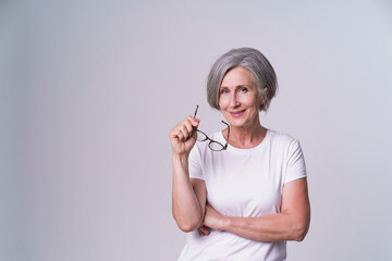 Mature businesswoman took off her glasses with folded hands looking straight in camera and smile wearing white t-shirt isolated on white background. Mature people beauty concept.