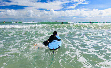 Young boy, a beginner in surfing, holding softboard and trying to bring it back into sea to...
