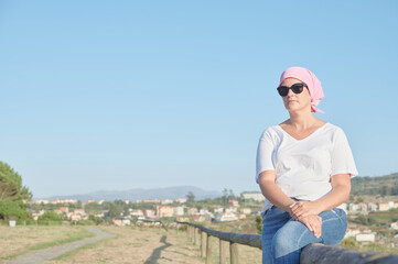 Fototapeta na wymiar World breast cancer awareness day October 13: Middle-aged woman suffering from cancer wears a pink scarf covering her head due to chemotherapy while looking at the ocean.