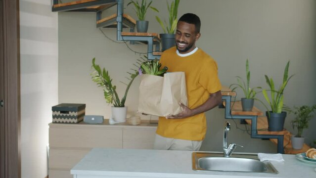 Slow motion of smiling African American man carrying bag of groceries walking in modern kitchen at home. Shopping and cooking concept.