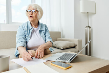 Portrait of an elderly business woman taking notes in a notebook sitting on the couch at home with a laptop undergoing online training