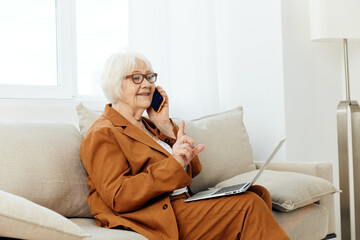 a pleasant, emotional old lady is sitting on a sofa in a beautiful apartment in a stylish brown suit talking on the phone with a laptop on her lap, holding her index finger up