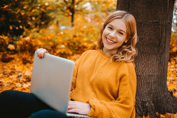 A beautiful girl teenager sits in an autumn park with a laptop. The student studies on the street. Study in the fall. The woman looks into the camera