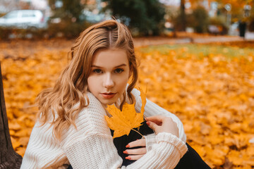 A girl in a white sweater sits on the autumn leaves. A beautiful blonde with a square looks at the camera. Woman outdoors in the autumn park