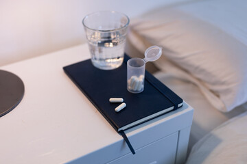 Close up of a notebook, box of tablets and a glass of water lying on a cabinet next to a bed
