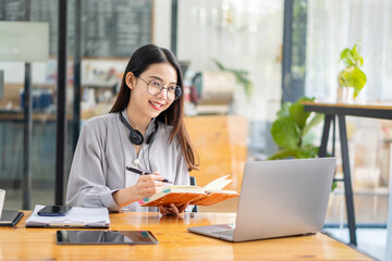 Fototapeta na wymiar Pretty Asian woman and headphones using laptop in coffee shop writing notes. Attractive female student learning language. View webinars online. audio course education