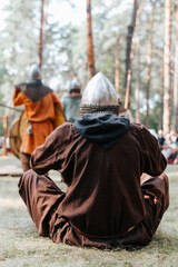 Medieval soldier in a helmet and ancient clothes sitting on the grass in the forest, rear view...