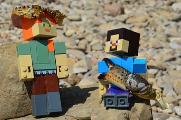 Fototapeta premium LEGO Minecraft figures of Steve and Alex on shoreline rock, Alex standing with crab cephalothorax on her head and Steve sitting with with crab arm and claw. Summer daylight sunshine.